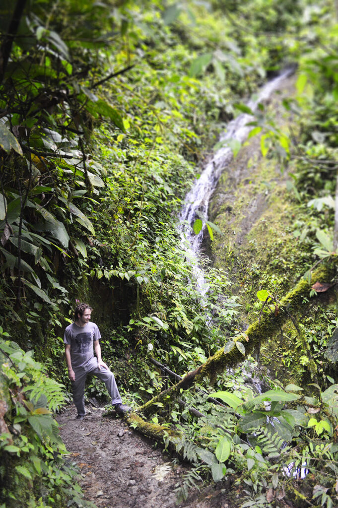 A man stands at the base of a small waterfall in a lush green Ecuadorian forest