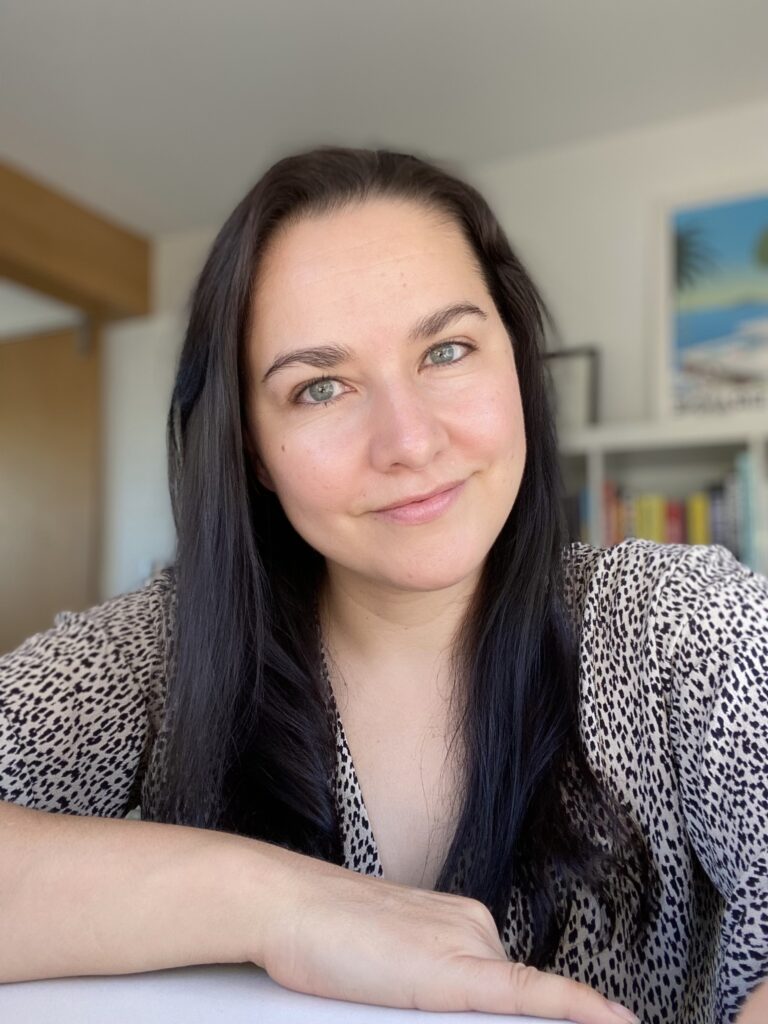 Portrait of a Latinx woman with olive skin, green eyes, black hair, leaning on a desk and looking at the camera with art displayed in background